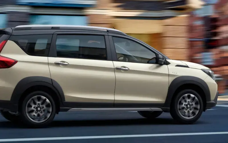 Suzuki XL7 Híbrida en movimiento, vista trasera de un vehículo familiar de color beige con detalles negros, recorriendo una calle urbana.