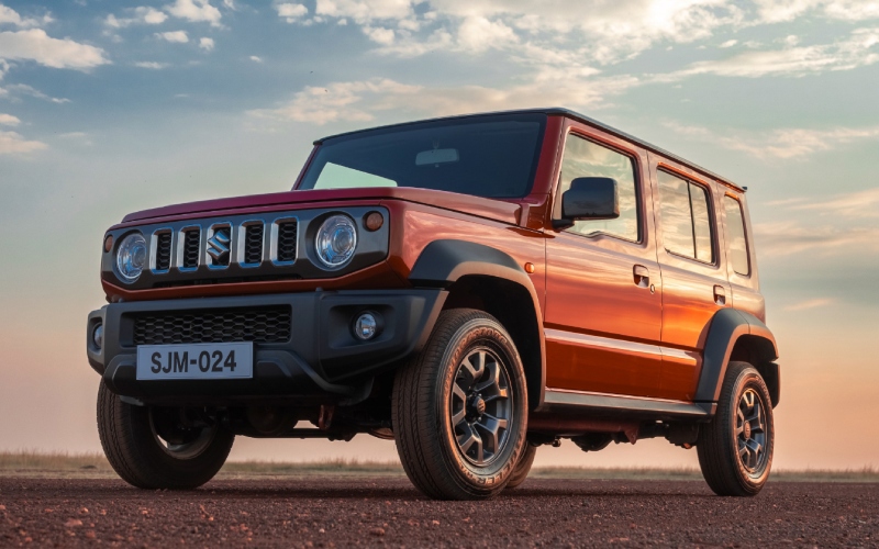 Suzuki Jimny 5 puertas de color rojo, con la parrilla delantera y los faros encendidos, estacionado en un camino de tierra durante el atardecer.
